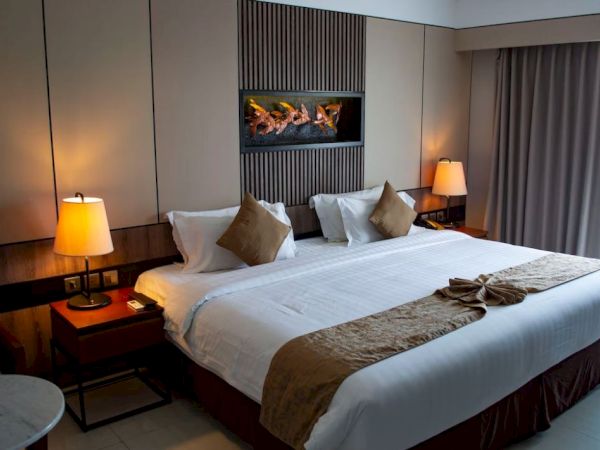 A modern hotel room with a neatly made bed, bedside lamps, and a lit fireplace feature on the wall.