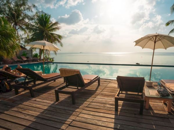 Beach resort scene with wooden deck, lounge chairs, umbrellas, and calm ocean under a cloudy sky.