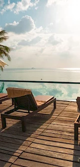 A beachfront resort deck with lounge chairs and umbrellas overlooking calm ocean waters under a bright cloudy sky.