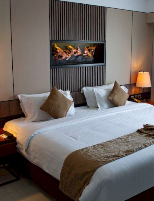 A modern hotel room with a neatly made bed, warm lamps, and a glowing firebox fireplace behind pillows.