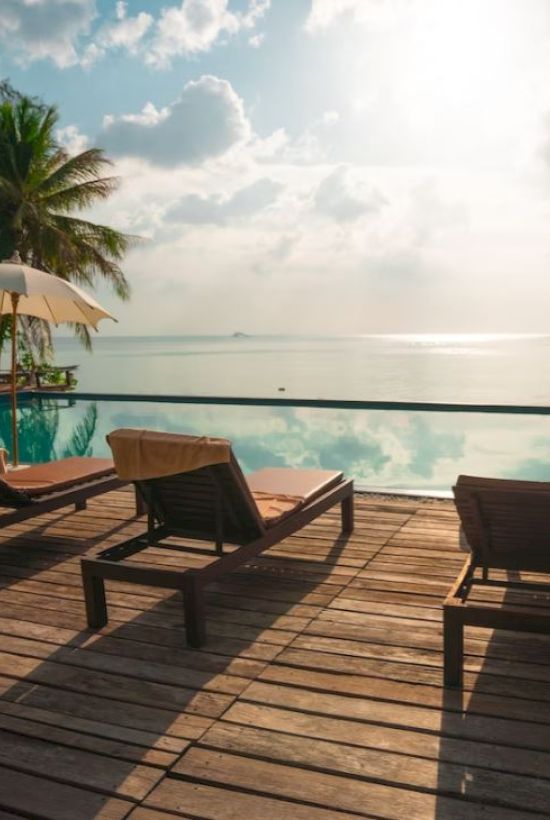 A tropical beachfront resort: wooden deck with lounge chairs, umbrellas, and a calm ocean at sunset.