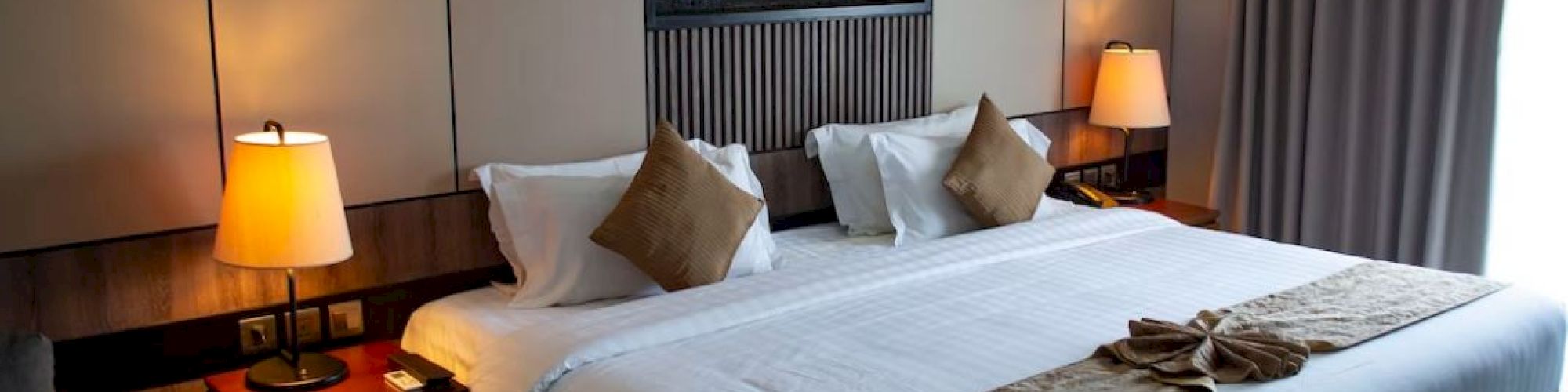 A modern hotel room with a neatly made bed, lamps, and a wall fireplace with flames.