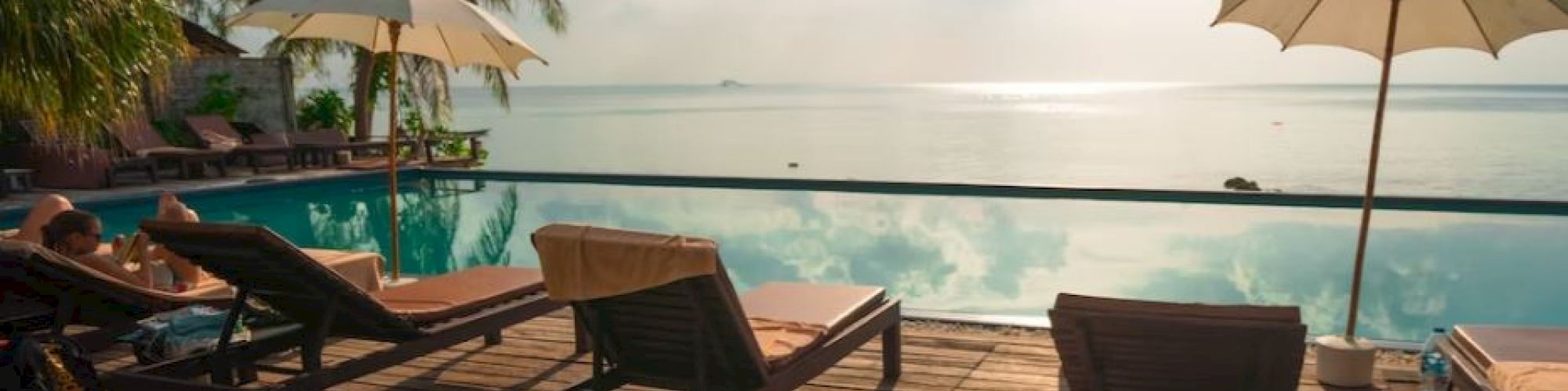 An oceanfront resort deck with lounge chairs, umbrellas, a pool, and palm trees under a cloudy sky.