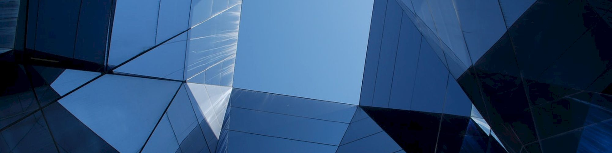 A geometric, blue glass skyscraper facade with sharp angular panels and reflections, shot upward into the sky.