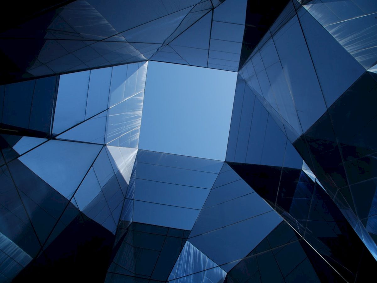 Angled blue glass architectural facade with geometric panels and reflections, like a modern high-rise viewed from inside.