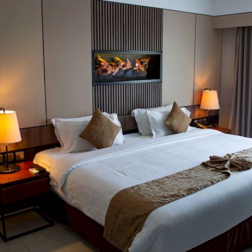 A neatly made hotel room with a large bed, bedside lamps, and a wall fireplace showing burning logs.