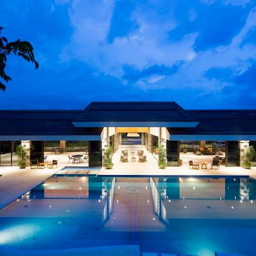 A modern hotel courtyard with a lit swimming pool under a blue evening sky and lounge chairs.