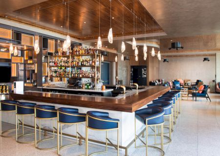 A modern hotel bar with a wooden counter, glass pendant lights, shelves of liquor bottles, and lounge seating in the background.