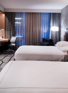 A modern hotel room with two neatly made beds, a desk chair, curtains, and a TV on the wall.