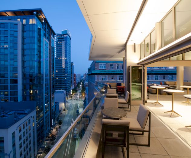 A high-rise city street view from a modern hotel balcony, with outdoor seating and warm lights fading into dusk.