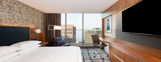 Modern hotel room with a bed, desk chair, TVs, wood paneling, and a window view of city buildings.
