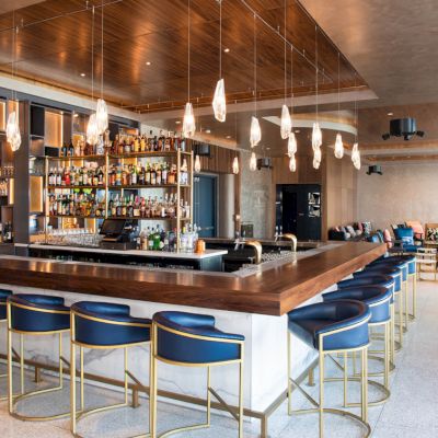 An upscale bar with a wooden counter, stocked bottle shelves, blue stools, pendant lights, and a lounge seating area.
