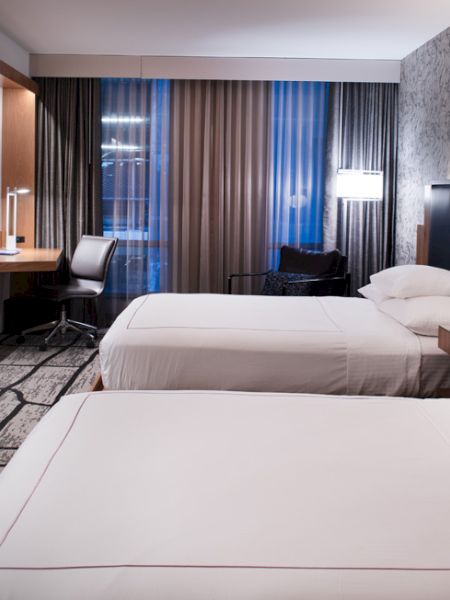 A modern hotel room with twin beds, wall-mounted TV, and striped curtains behind a seating area.