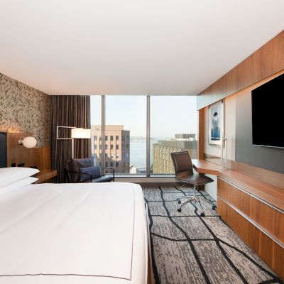 A modern hotel room with a large bed, desk chair, built-in TVs, and floor-to-ceiling windows showing a city view.
