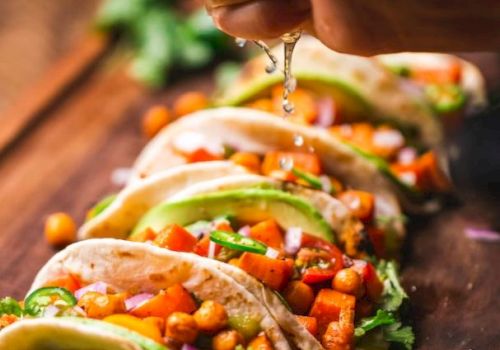 A person bites into tacos, letting melted sauce drip over a filling of veggies, beans, and cilantro, with lime nearby.