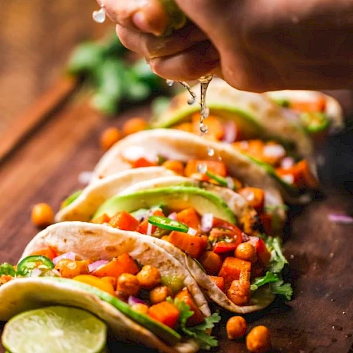 A person bites into tacos, letting melted sauce drip over a filling of veggies, beans, and cilantro, with lime nearby.