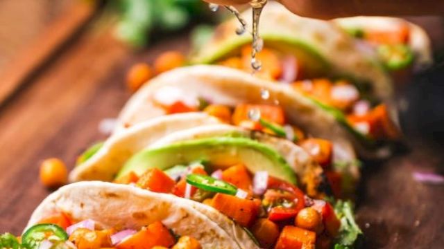 A person bites into tacos, letting melted sauce drip over a filling of veggies, beans, and cilantro, with lime nearby.