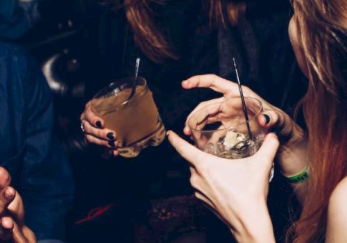 Two women with long nails and rings sip cocktails in a dim night club, each holding a drink with a straw and ice.