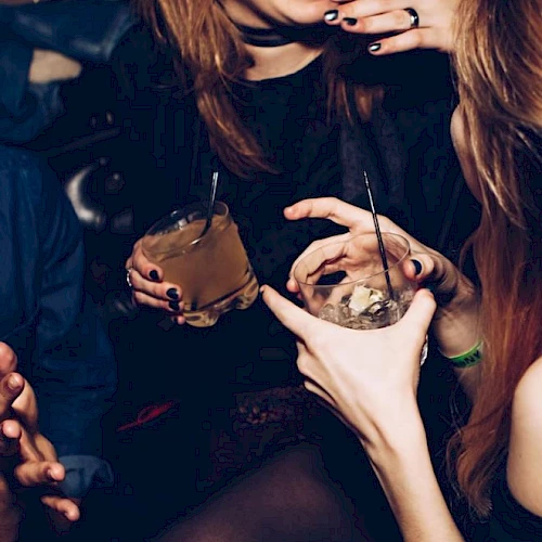 Two women with long nails and rings sip cocktails in a dim night club, each holding a drink with a straw and ice.