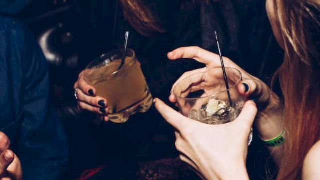 Two women with long nails and rings sip cocktails in a dim night club, each holding a drink with a straw and ice.