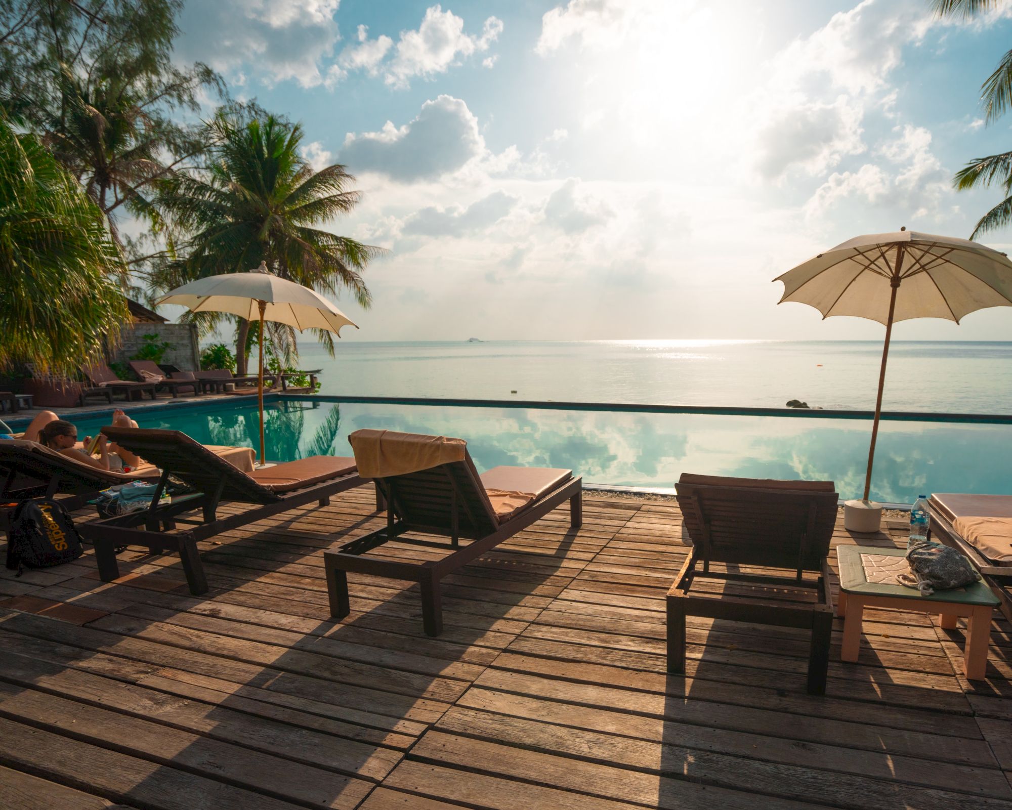 A tropical beach resort deck with loungers, umbrellas, a pool, and ocean views under sunny clouds.