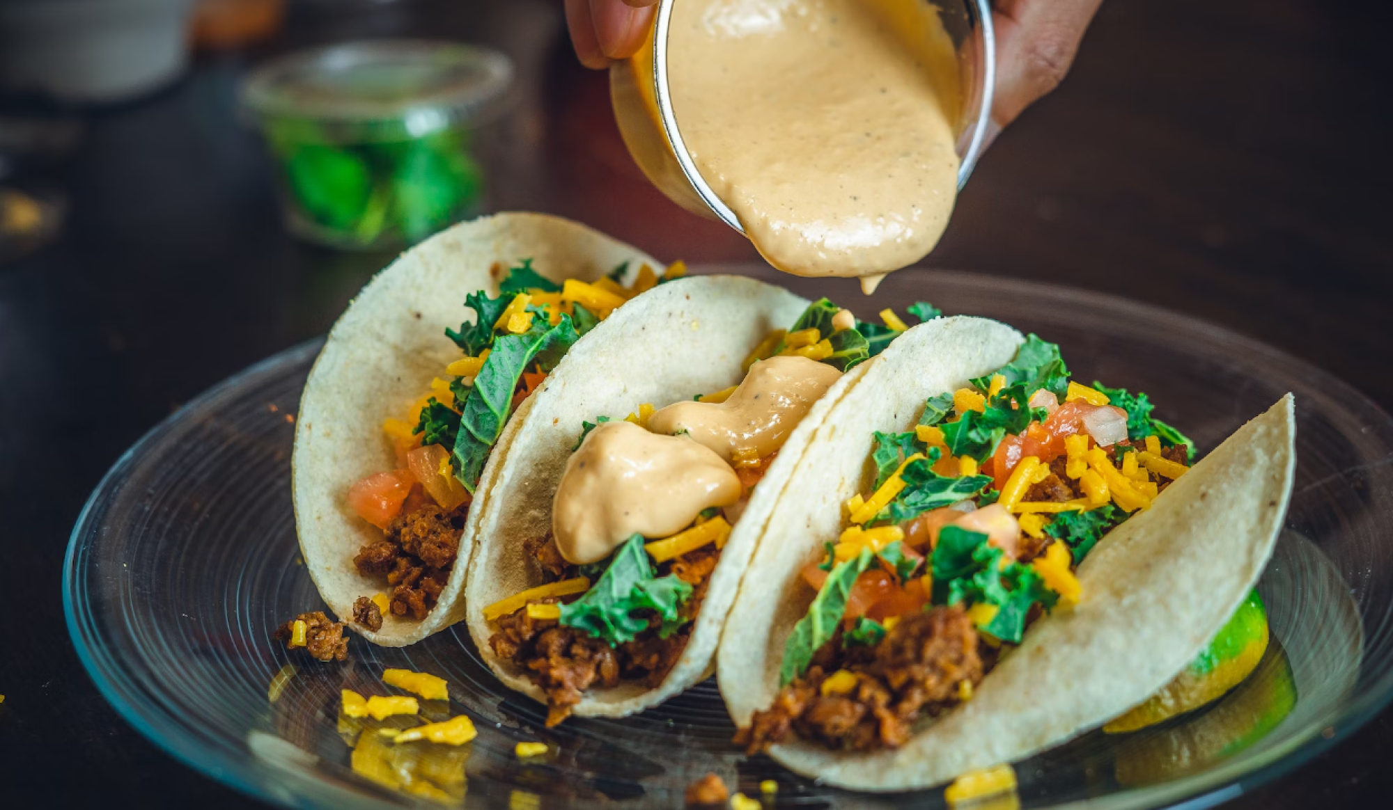 A hand pours creamy sauce onto three tacos filled with seasoned meat, shredded cheese, and chopped herbs on a plate.