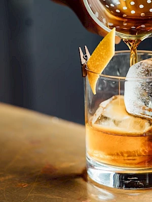 A cocktail being poured into a short glass with ice, featuring a garnish of an orange slice and a toothpick.