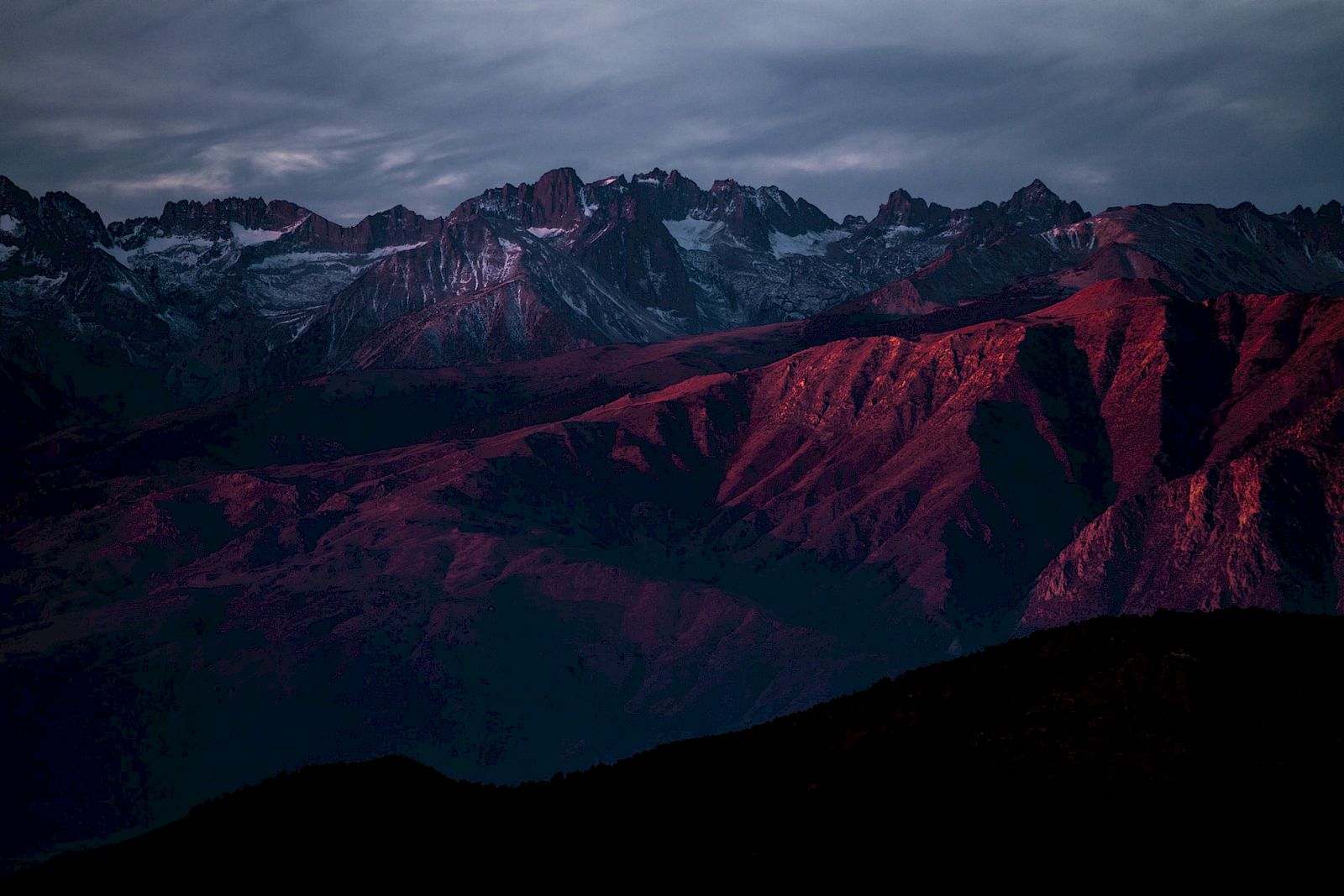 Dramatic mountain range at dusk with snowy peaks and red-lit slopes under stormy clouds, like a rugged alpine landscape.