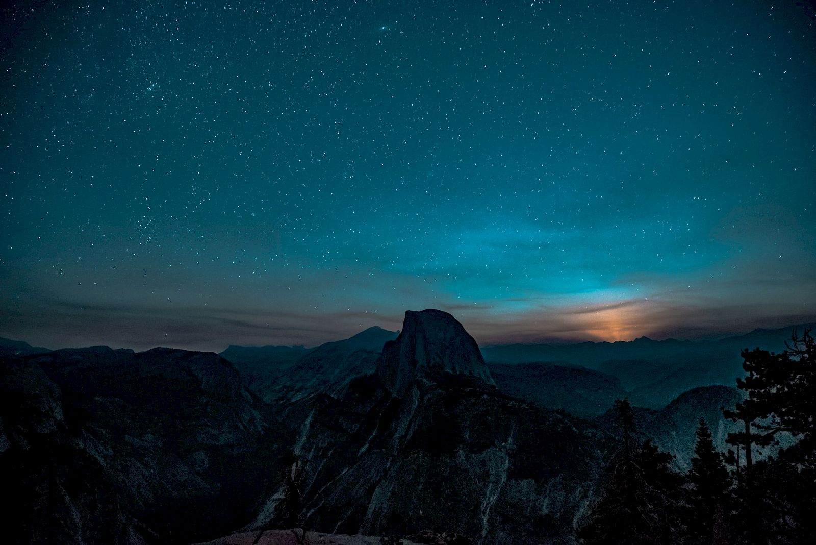 A star-filled night sky glows above dark mountains, with a bright, misty horizon and a towering rocky peak below.