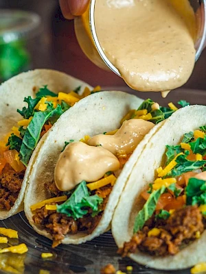 Three tacos on a plate topped with chopped veggies and shredded meat, with creamy sauce being drizzled on top.