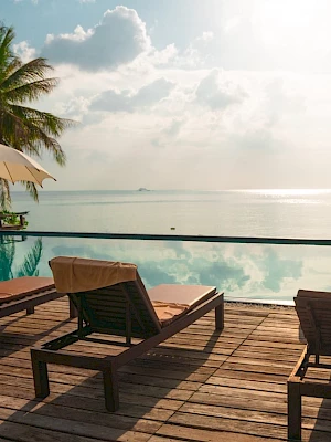 A tropical beachfront resort deck with a pool, sun loungers, umbrellas, and palm trees overlooking calm ocean at sunset.
