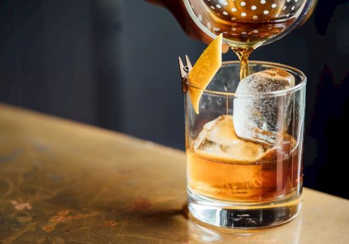 A bar pours amber liquor into a rocks glass with ice, topped with a skewered citrus peel garnish.