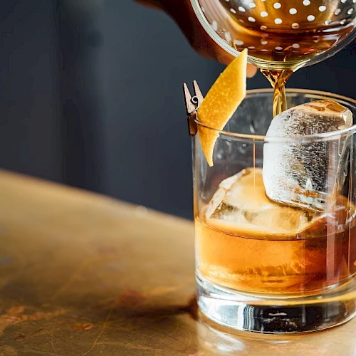 A bar pours amber liquor into a rocks glass with ice, topped with a skewered citrus peel garnish.