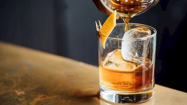 A bar pours amber liquor into a rocks glass with ice, topped with a skewered citrus peel garnish.