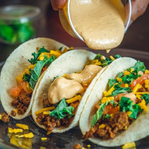 Three tacos on a plate, topped with shredded lettuce, diced tomatoes, cheese, and creamy sauce being poured over them.