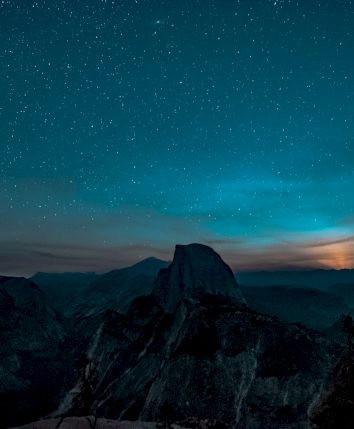 A star-filled night sky over dark mountain peaks, with a glowing horizon and wisps of clouds drifting across the valley.