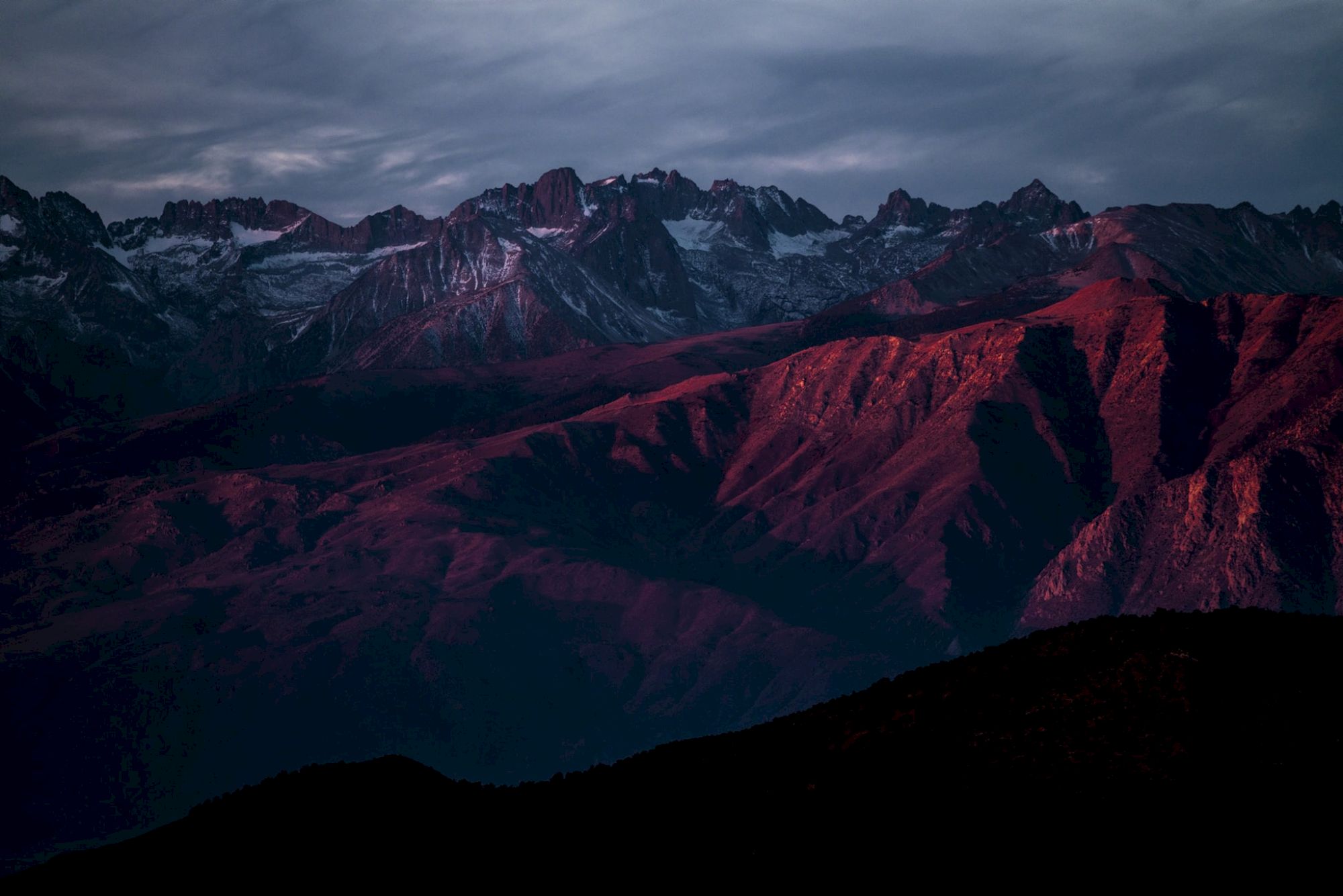 Breathtaking mountain range at dusk, with snow-capped peaks and crimson-lit ridges under a stormy sky.