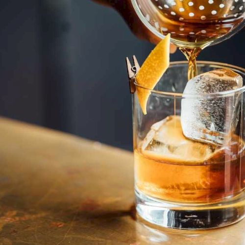 A whiskey glass with a large ice cube, a citrus twist garnish, and a small chip clip perched on the rim, poured with amber liquid.