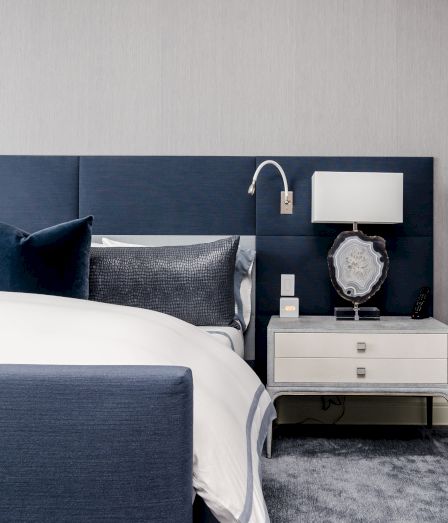 A modern blue hotel bedroom with a made bed, nightstand, lamp, remote, clock, and a decorative stone on the dresser.