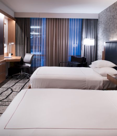 A modern hotel room with two neatly made beds, a flat-screen TV, wood paneling, and tall curtains framing a window.