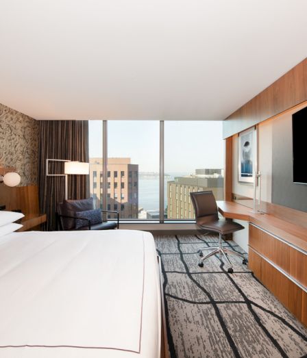 A modern hotel room with a large bed, desk chair, TVs, and floor-to-ceiling windows overlooking buildings.