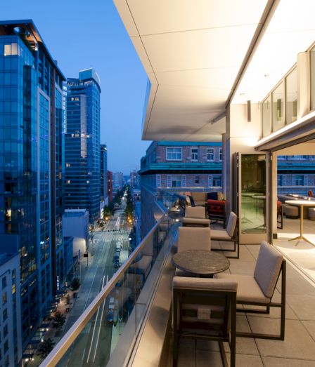 A rooftop balcony in a high-rise overlooks a downtown street between glass offices, with outdoor seating and bright daylight.