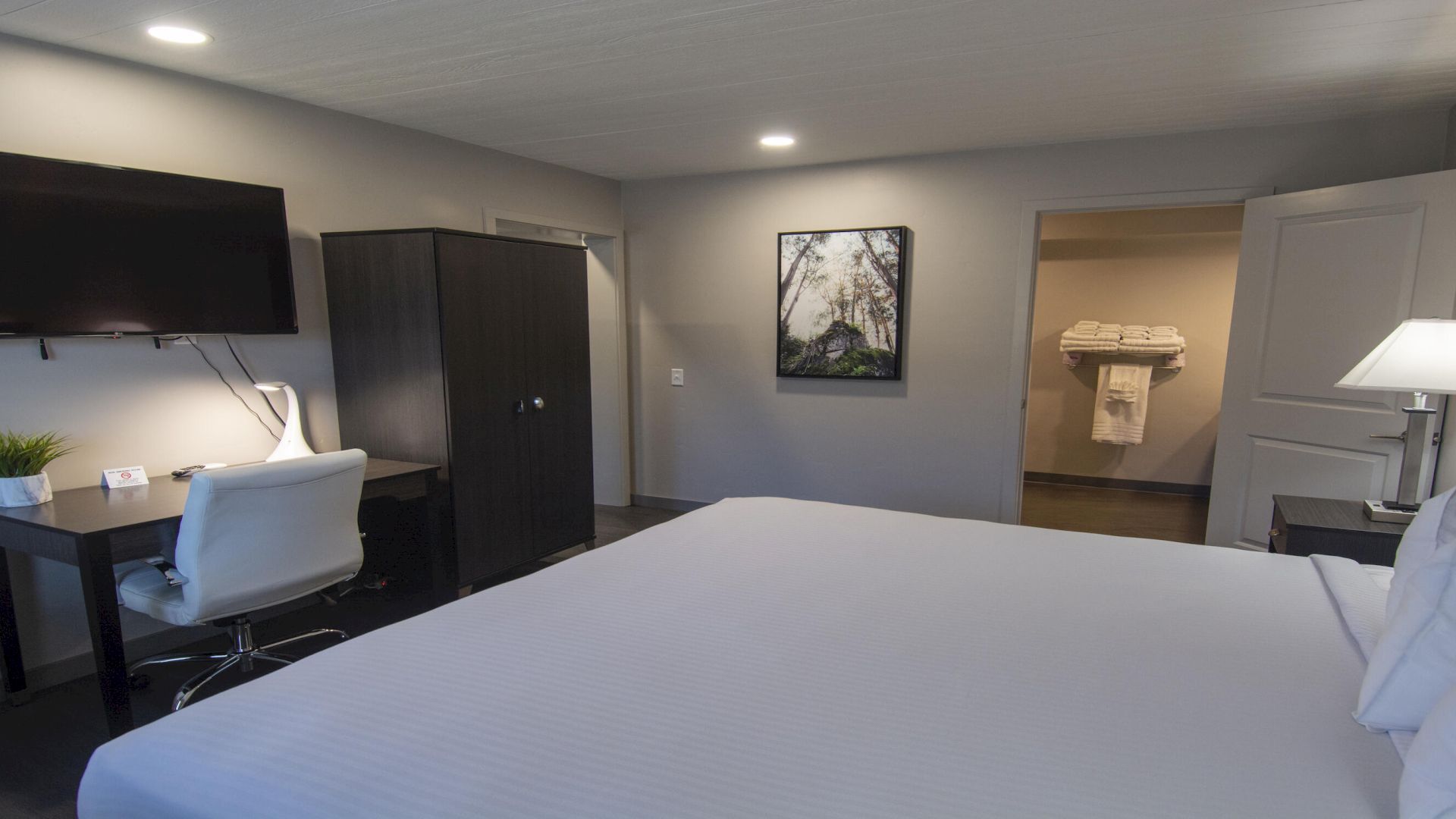 A modern hotel room with a large bed, a desk and chair, a mounted TV, a wardrobe, and a view into a bathroom with a towel rack.