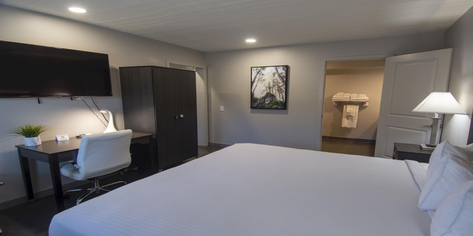 A modern hotel room with a large bed, a desk and chair, a mounted TV, a wardrobe, and a view into a bathroom with a towel rack.