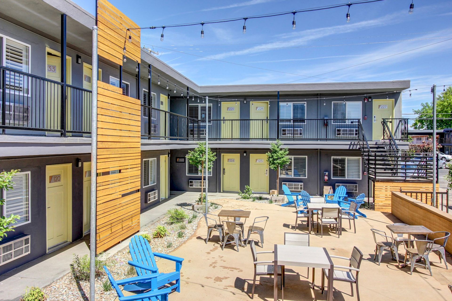 Court-style apartment complex courtyard with string lights, patio tables, and colorful chairs; two-story gray buildings with yellow accents.