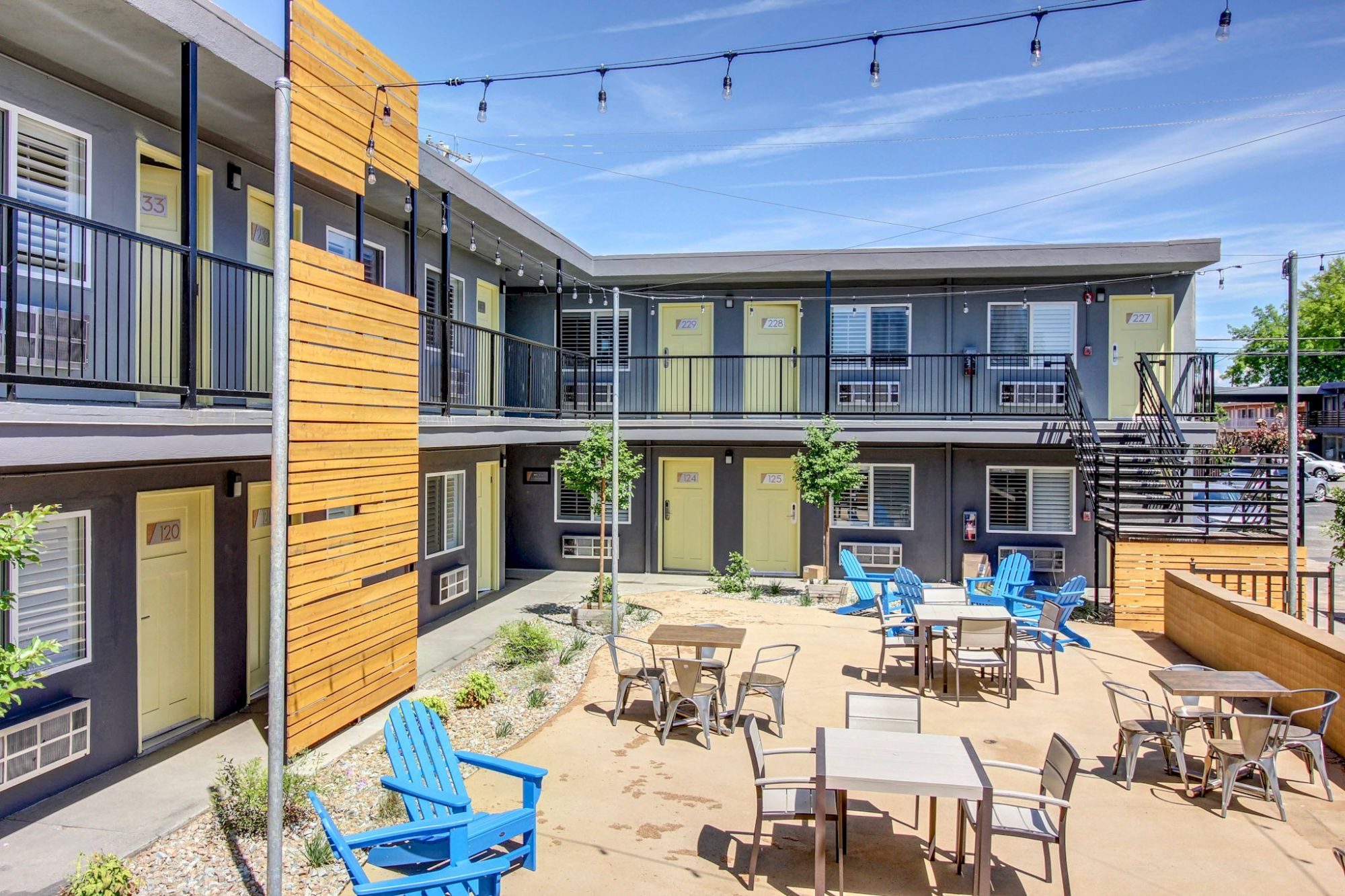 Court-style apartment complex courtyard with string lights, patio tables, and colorful chairs; two-story gray buildings with yellow accents.
