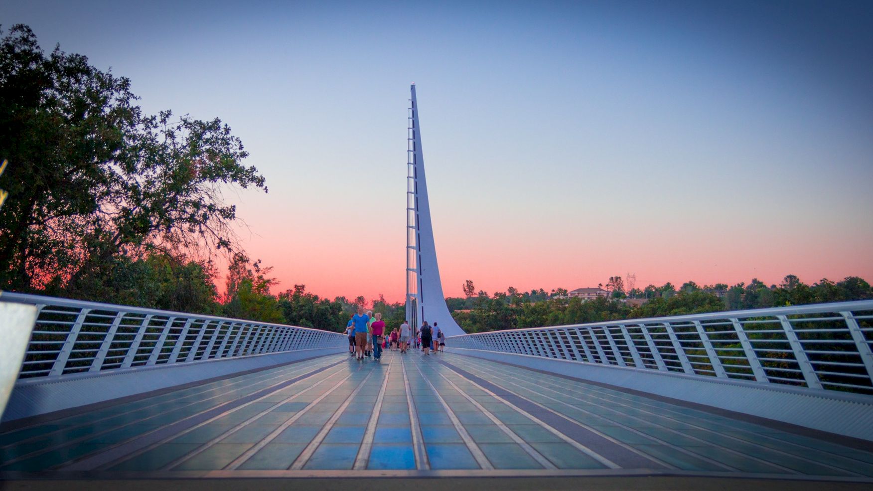 People walk on a modern bridge at sunset as a tall tower rises against the sky.