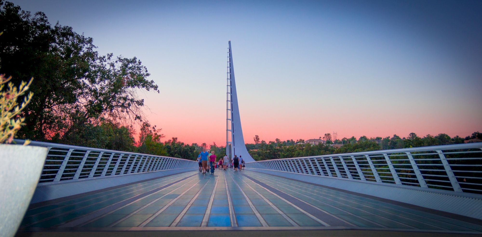 People walk on a modern bridge at sunset as a tall tower rises against the sky.