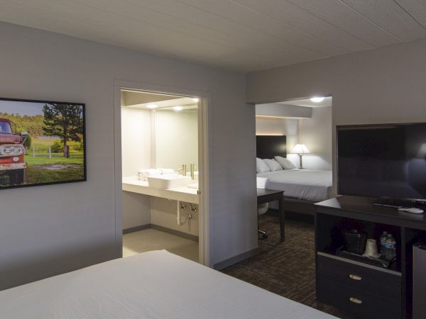 A hotel room with a bed in foreground, a doorway to a bathroom, a TV and cabinet opposite, and a wall-mounted framed truck photo.