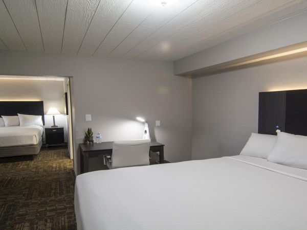 A modern hotel suite with a large white bed in the foreground, a desk and chair, and a separate bedroom visible through an open doorway.